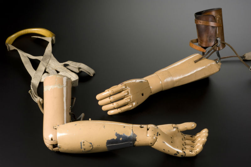 Left above elbow prosthesis with a wooden socket connected to a wooden forearm. Metal mechanical hand with passive flexion and extension of the wrist. Elbow flexion is combined with pronation / supination of the wrist. Made by Carnes Arm Company, United States of America 1915. Left below elbow prosthesis with a wooden socket, connected by jointed side steels to a leather upper arm corset. Metal mechanical hand with pronation / supination action combined with flexion and extension of the elbow. Hole in hand for driving cup fitment (added in more recent years) Passive wrist flexion and extension. Made by Carnes Arm Company, United States of America 1915. Graduated matt black perspex background. c. Science Museum, SSPL