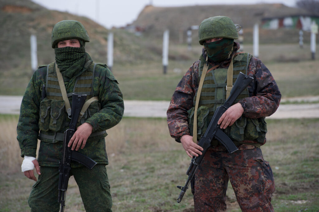 Two Russian soldiers wearing green without insignia, both wearing helmets, face masks, and black rifles. 