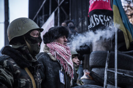 Two Ukrainian protestors stand at a tyre barricade. One is wearing a camouflage jacket, balaclava and military helmet. The other is wearing a dark jacket and a black hat.