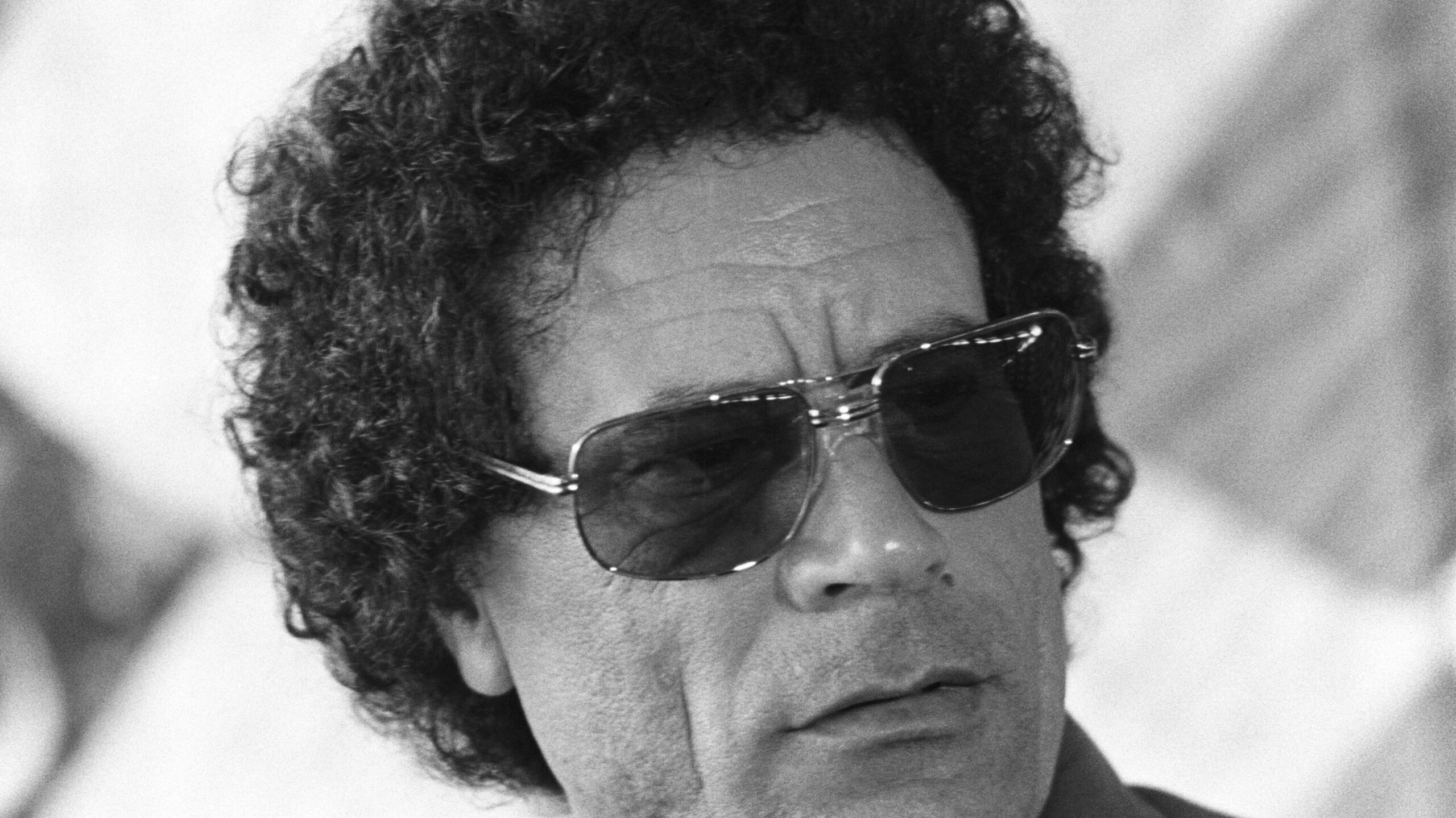 Colonel Muammar Gaddafi in black and white wearing sunglasses.