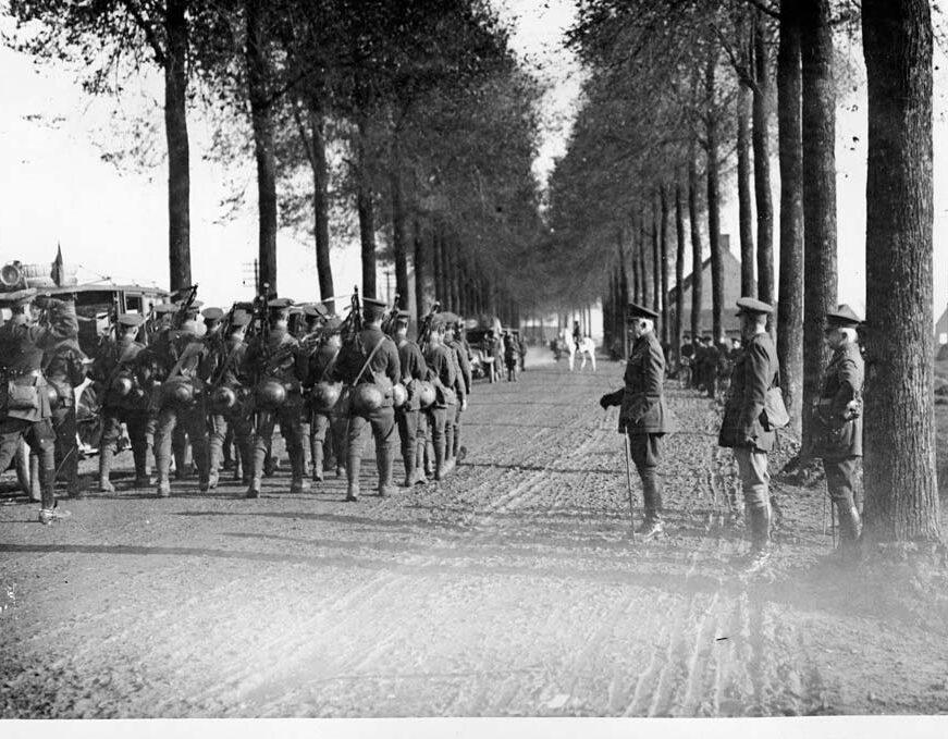 Visiting dignitaries watch on as infantry march past along a tree-lined dirt road. 