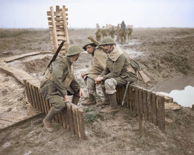A colourised photograph showing three exhausted men sitting on a fence in a muddy landscape filled with shell holes. 