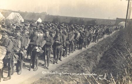 Hundreds of soldiers carrying rifles march past along a rural English road.