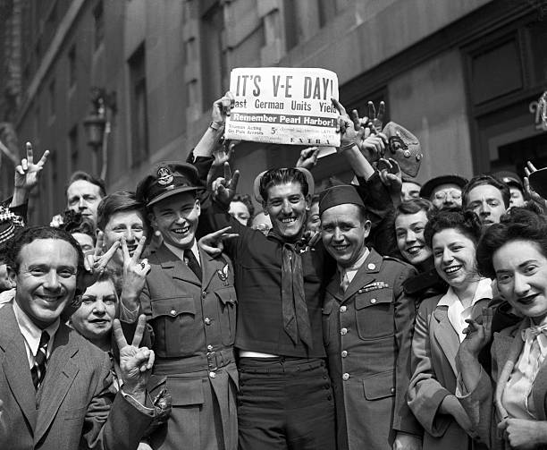 A crowd of men and women, including two uniformed servicemen, face the camera and smile. Many make 'V' signs with their fingers and a man in the middle holds up a newspaper with the headline: "it's V-E Day!