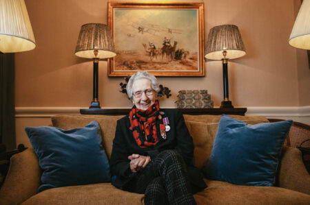 Marie Scott, wearing glasses, a red scarf, a black jacket, and her military medals sits on a sofa with two cushions. Behind is a painting showing WWI-era aircraft.