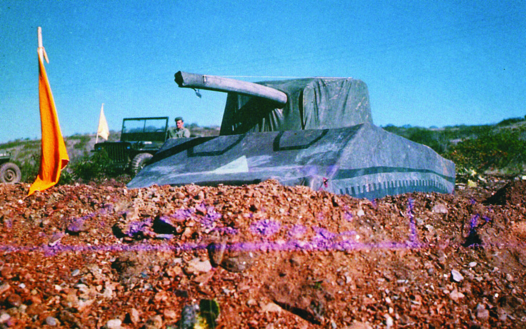 A colour image of an inflatable tank in a field against a blue sky. A soldier and a jeep are behind.